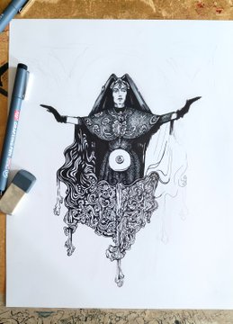 An intricate black ink drawing on paper depicts a hooded figure with outstretched arms, wearing an ornately patterned dress, surrounded by art supplies on a wooden desk.