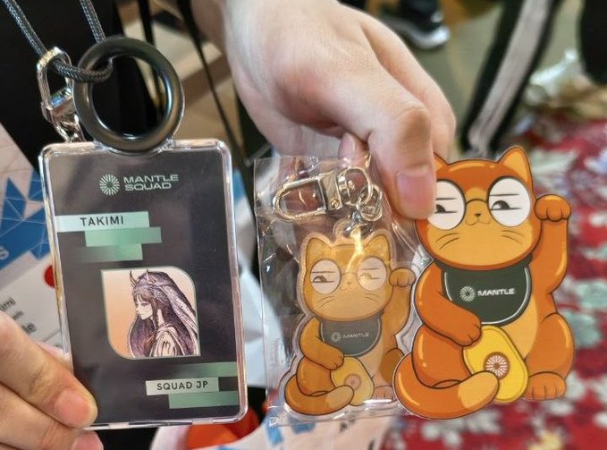A person holding Mantle Squad merchandise, including a Takimi ID card holder and acrylic keychains featuring cat characters wearing Mantle-branded shirts.
