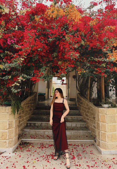 A woman in a burgundy dress stands beneath a vibrant archway of red and orange bougainvillea blooms, perfectly capturing the essence of #FlowersOnFriday. The cascade of fallen petals at her feet adds a romantic touch to this floral Friday scene.
