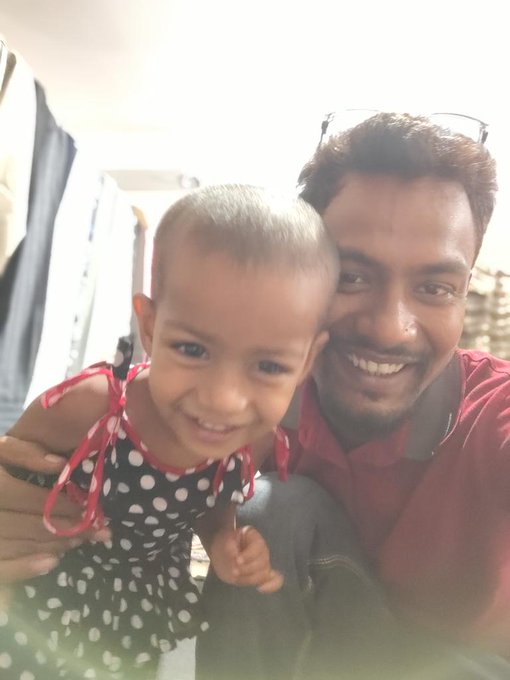 The image features a man sitting next to a young child who is wearing a dress with black polka dots. They are both smiling for the camera, creating a heartwarming scene. The background of the photo shows a room with a window that allows natural light into the space. There is also a chair visible in the background.