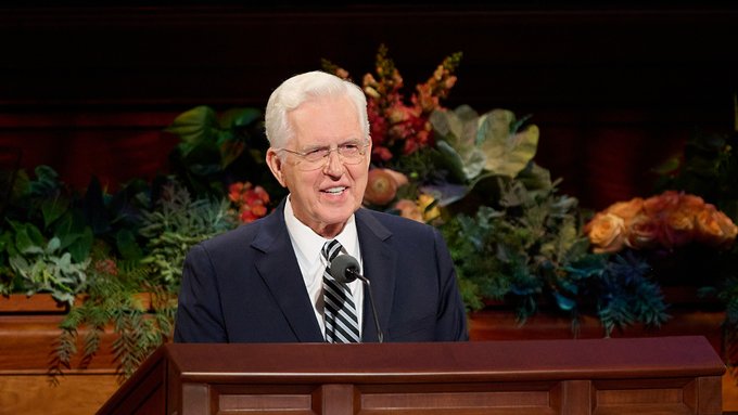 Portrait photograph of an older gentleman with white hair, glasses, and a dark suit smiling warmly at a wooden podium with a microphone, set against a backdrop of autumn floral arrangements.
