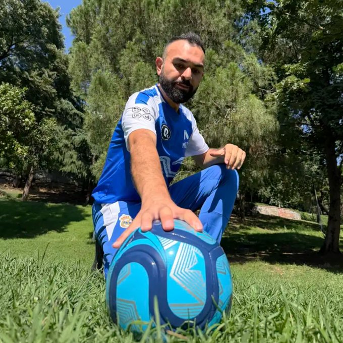 A bearded man in a blue and white sports uniform kneels on grass, holding a blue and black soccer ball, with trees in the background.