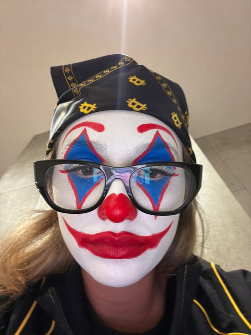 A person wearing dramatic clown makeup with white face paint, red lips, a red nose, and blue diamond designs around the eyes, black-framed glasses, and a dark bandana with Bitcoin (₿) logos — appearing to smile wryly at a self-portrait. The accompanying post jokes that people who ignored her work suddenly reach out only to ask for a favor (specifically a connection to @bcgame), with the clown imagery humorously conveying her disbelief and frustration at the situation.