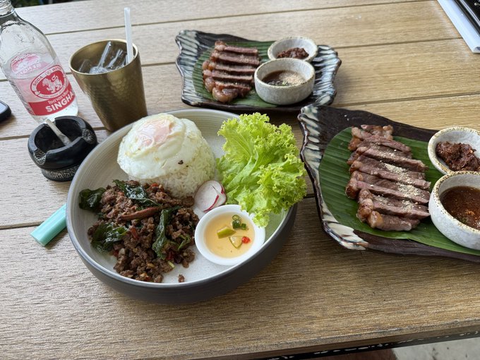 A Thai meal is spread on a wooden table, featuring rice topped with a fried egg, stir-fried beef with basil, fresh greens, and grilled pork with dipping sauces, accompanied by a Singha water bottle.
