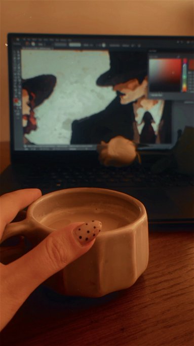 A hand with polka-dotted nail polish holds a beige ceramic mug of coffee in front of a laptop screen displaying digital art of a figure with a hat and mustache.