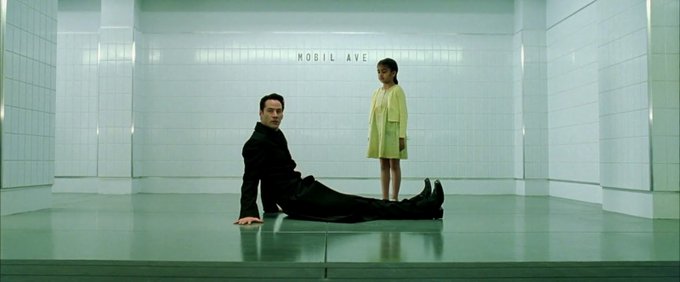 A man in a black suit sits on the floor of a sterile, green-tiled waiting room labeled "MOBIL AVE," while a young girl in a yellow dress stands nearby, evoking the retro-futuristic aesthetic reminiscent of sci-fi waiting areas. The cold, minimalist environment aligns with the "@zaddrnet" post's reference to a "zaddr approval waiting room," suggesting a dystopian or otherworldly bureaucratic setting.