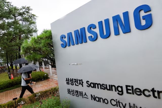 A pedestrian with an umbrella walks past a Samsung Electronics sign at the company's Nano City facility in Suwon, South Korea, on a rainy day. The image visually anchors the $SAMSUNG stock discussed in the accompanying post about Q2 earnings preview and 24/7 perpetual futures trading on Decibel.
