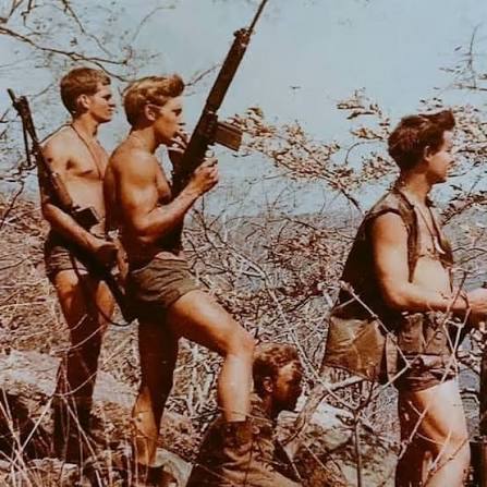 A vintage photograph depicts several shirtless young men in shorts carrying rifles as they walk through a brushy, outdoor environment.