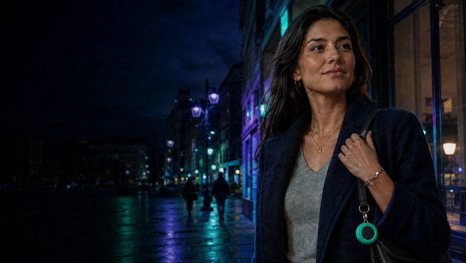 A woman with long dark hair wearing a gray top, dark coat, and a green circular pendant, standing on a city street at night with illuminated buildings in the background.