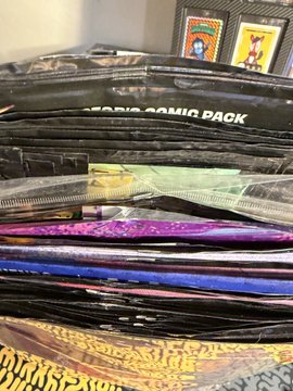 A stack of glossy black packages, with one labeled 'PARIS COMIC PACK' and another showing 'STICKERS' and 'TOPPS' text, alongside colorful metallic foil packs.