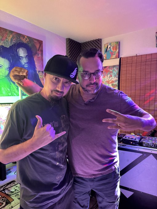 Two men posing together indoors with purple and pink lighting, one wearing a black cap and t-shirt and the other in a purple v-neck shirt and glasses, standing in front of artwork and DJ equipment.