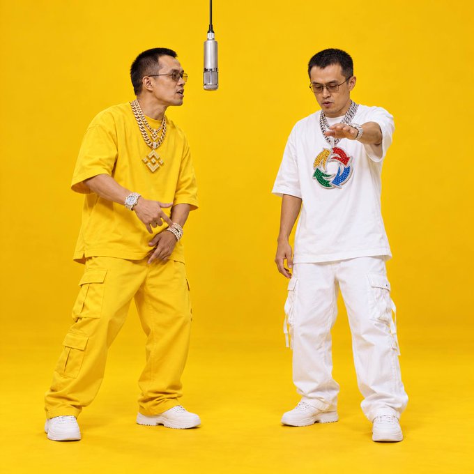 Two men stand against a yellow background with a microphone hanging between them; the man on the left wears all yellow with large gold chains, while the man on the right wears a white t-shirt with a colorful design and white cargo pants.
