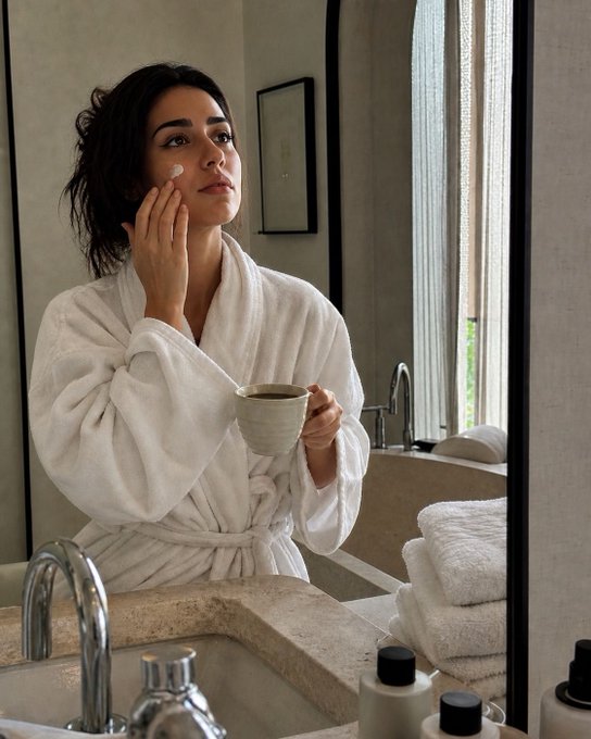 A woman with wet, curly dark hair, wrapped in a white bathrobe, stands in a bathroom applying cream to her face while holding a small bowl and looking in the mirror.