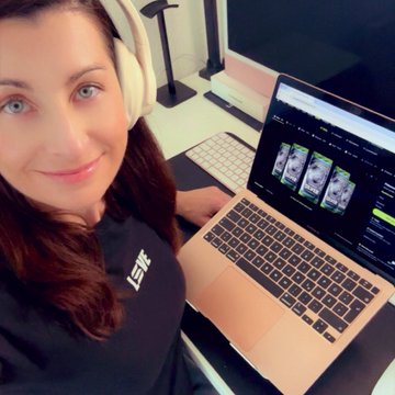 A selfie of a smiling woman wearing headphones, sitting at a desk with a laptop displaying video editing software.