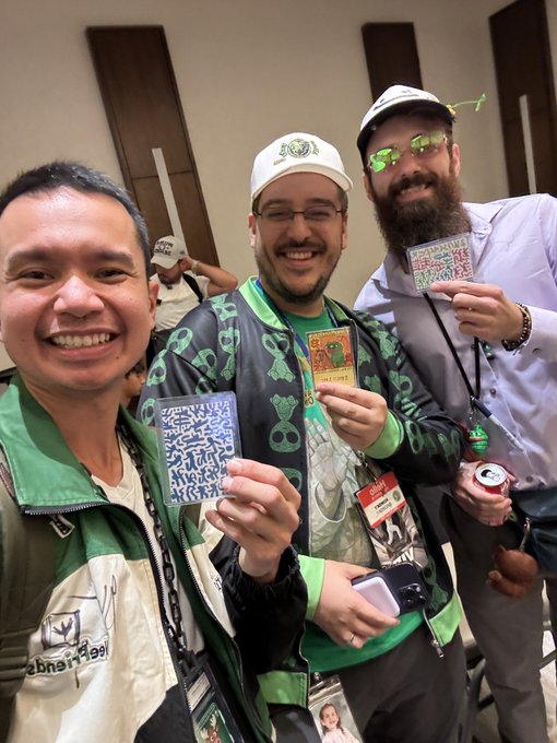 Three men smile for a selfie while holding up their convention badges at what appears to be a gaming or trading card event, with lanyards visible.