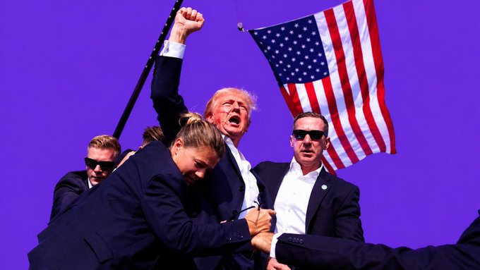 Donald Trump appears in a rally scene holding an American flag while surrounded by security personnel, including an agent restraining a protester.
