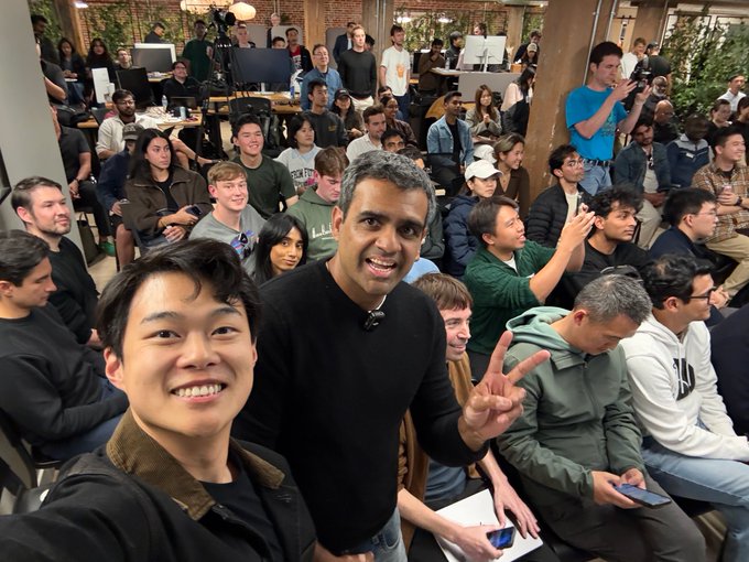 Two men take a cheerful selfie in the foreground—one in a black sweater flashing a peace sign and another in a brown corduroy-collared jacket—while a large audience of seated attendees fills the background, suggesting a lively tech or hacking event hosted with @TheHumanoidHub and @hackersquadio.