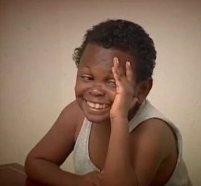 A vintage photograph of a smiling young boy with his hand pressed against his face in a playful gesture.