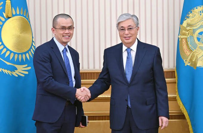 A formal photograph of two men in dark suits shaking hands in front of the Kazakhstan flag (blue with a gold sun and eagle), appearing to be an official diplomatic meeting.