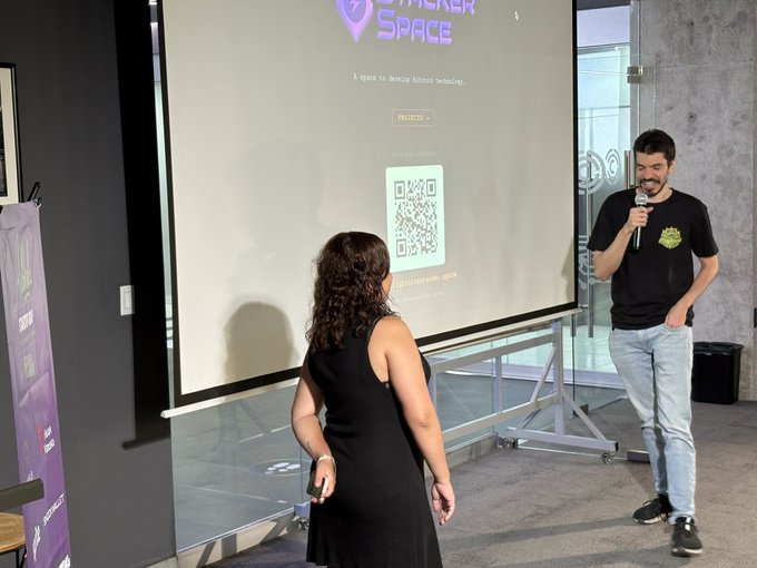 A man with a microphone speaks to a woman in a black dress at a 'Hacker Space' event, with a QR code displayed on the projector screen behind them.