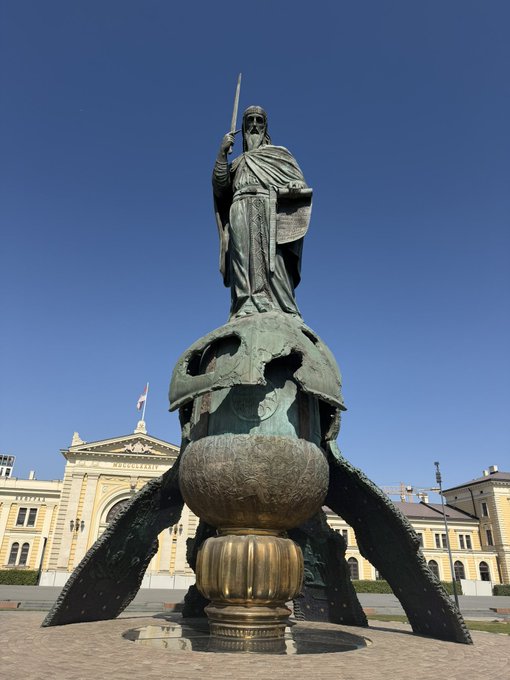 A bronze statue of a robed religious figure holding a sword and book, standing atop a decorative sphere pedestal, with classical buildings in the background under a clear blue sky.