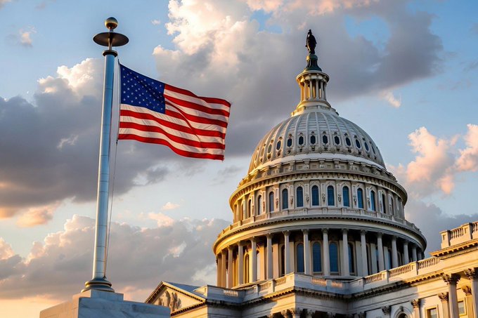 The U.S. Capitol building dome with a large American flag flying on a flagpole in the foreground, set against a partly cloudy sky with warm sunset lighting.