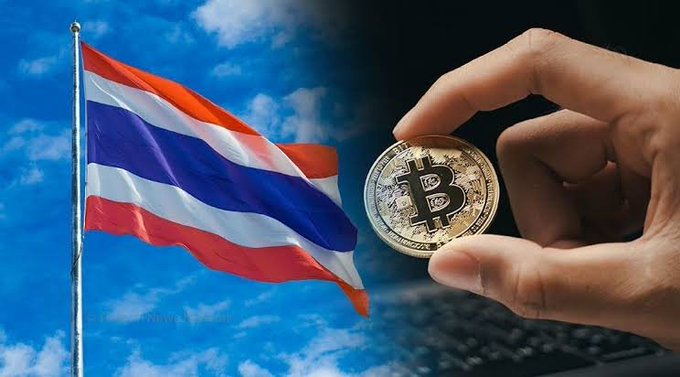 A hand holds a physical Bitcoin coin next to the waving flag of Thailand against a blue sky with clouds, with a keyboard visible in the background.