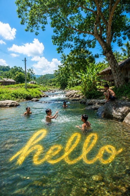 Children swim and play in a clear tropical river surrounded by lush greenery and a large tree, with the word 'hello' written in glowing yellow script over the water.