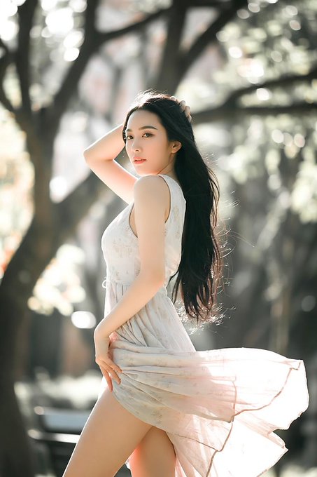 A young woman with long dark hair in a flowing white floral dress poses in a sunlit forest setting, lifting her hair and the hem of her dress.