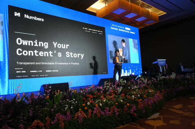 The image shows a large stage with a banner that says "Owning Your Content's Story" written in black text. A man stands behind the stage, presenting or speaking about content authenticity. The stage has flowers and plants surrounding it, creating an inviting atmosphere for the event. There are speakers visible on either side of the stage, indicating a well-organized presentation.