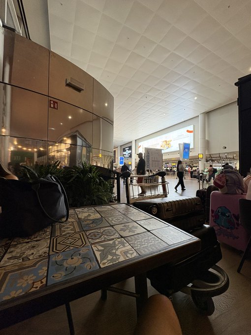 A photograph taken inside what appears to be an airport terminal, looking past a patterned tile table with a dark bag resting on it toward other travelers, luggage, and a baggage claim or gate area in the background.