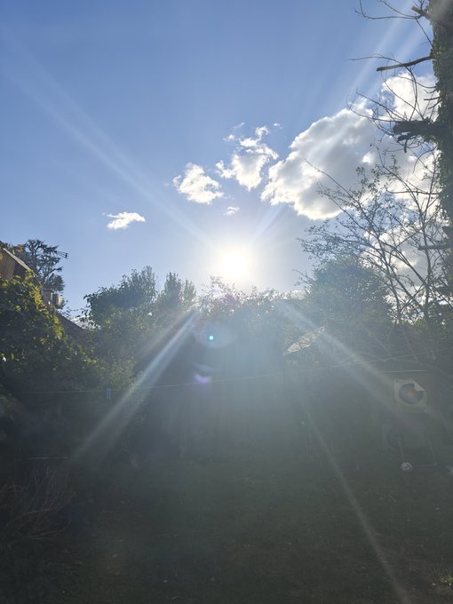 A bright sun creating prominent sun rays streaming through scattered clouds over trees and a backyard with a trampoline.