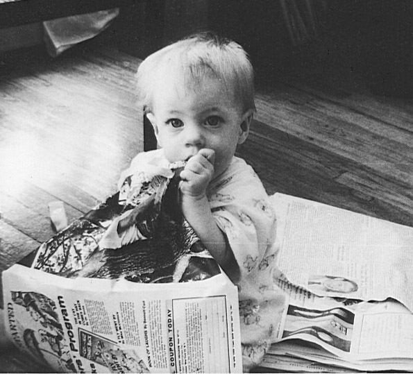 A black-and-white photograph of a young toddler sitting on newspaper, eating what appears to be a snack.