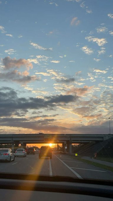 A view from inside a vehicle on a highway at sunset, showing cars driving under an overpass bridge with a colorful sky filled with clouds and the sun setting on the horizon.