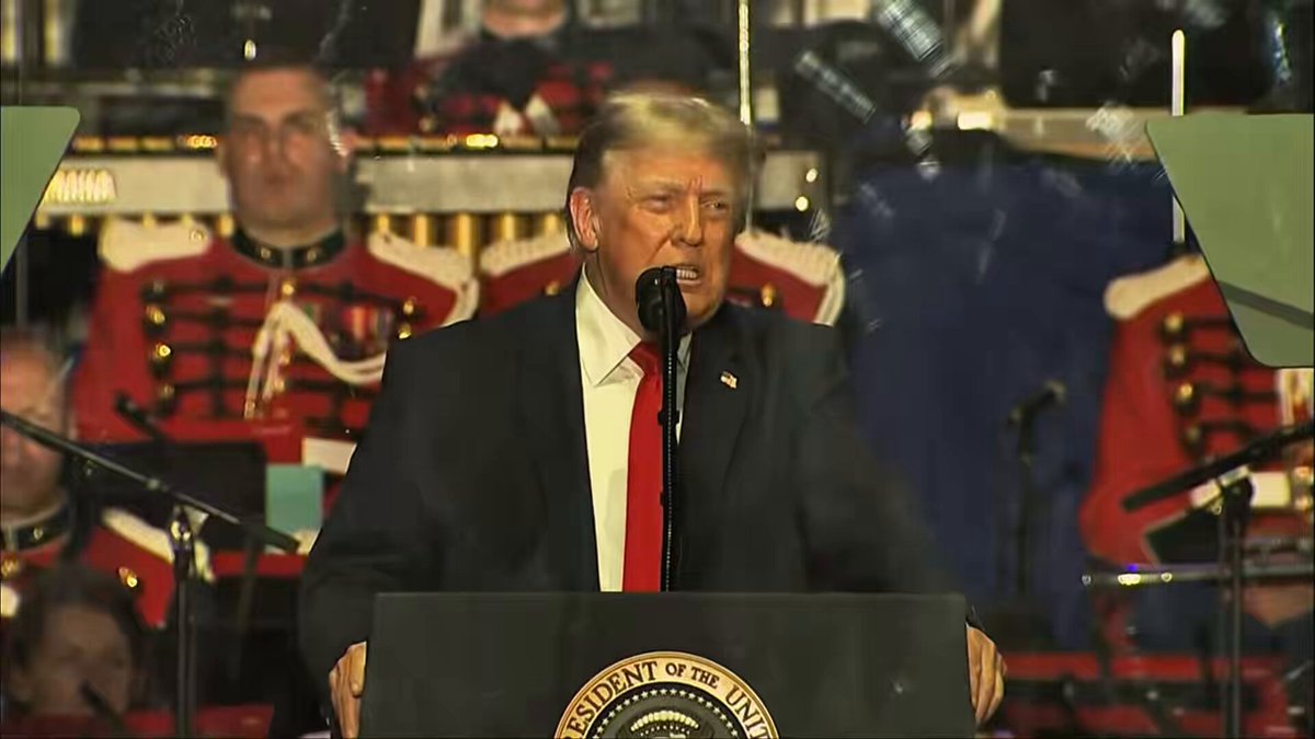 The image shows a man standing at a podium, giving a speech or addressing an audience. The man appears to be wearing a suit and tie, suggesting that he might be a political figure or leader. He is speaking into a microphone, which can be seen in front of him on stage. The background features several other people, indicating that this event may be taking place at a public gathering or conference.