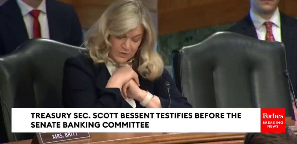 The image shows a woman sitting at a desk with her hands clasped together while she appears to be praying or deep in thought. The background of the image features a news conference setting, with a podium visible behind the woman. The text on the image reads "Treasury Sec., Scott Bessent calls for Congress to pass crypto market structure legislation before it'S too late." This suggests that the woman is addressing an important issue related to cryptocurrency and its regulation in the United States.