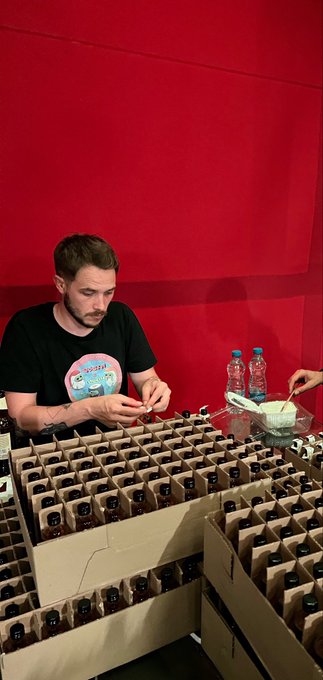 A man with a beard wearing a Squirtle t-shirt works at a table filled with numerous small bottles of what appears to be hot sauce, with a red backdrop behind him.