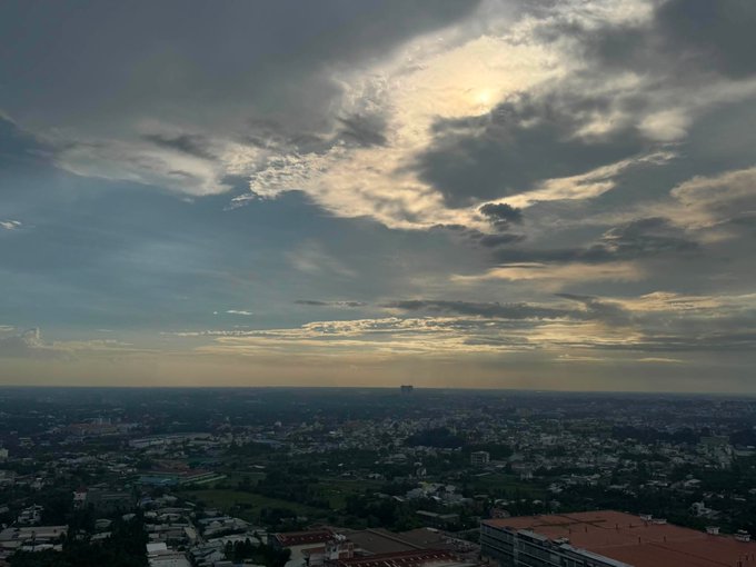 An aerial cityscape view shows a sprawling urban area under a dramatic sky filled with scattered clouds and sunlight breaking through.