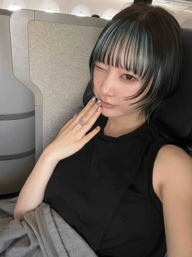 A young woman with dark green-tinted hair sits in what appears to be an airplane seat, wearing a black sleeveless top and showing a ring on her finger. The image complements the travel post about arriving in Tokyo and finding Tesla Superchargers at Narita Airport.