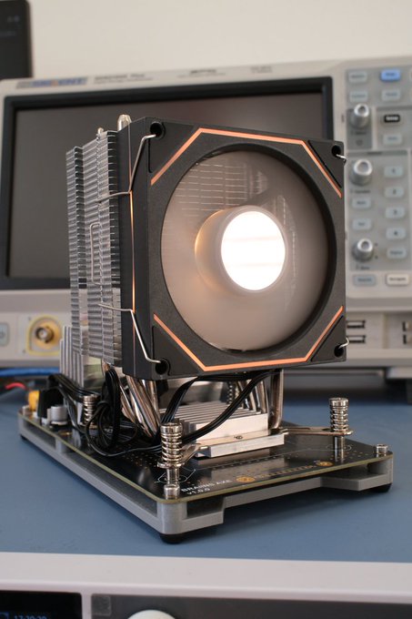 A photo of a large CPU cooler with a fan mounted on a test bench, positioned in front of an oscilloscope or testing equipment.