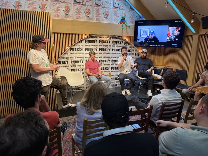 A panel discussion takes place at PubKey NYC, with the venue's logo and "WHAT'S COOKING ON BITCOIN?" text visible on the backdrop, as Bitcoin CEO Rob Ham, Matt Corallo (TheBlueMatt), and other Bitcoin developers address a packed, standing-room-only audience.