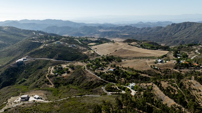 A sweeping aerial view captures a rugged mountain landscape with winding dirt roads, sparse homes, and rolling hills under a hazy sky. The post highlights this as a personal weekend photography shot, showcasing the scenic beauty of the elevated terrain.