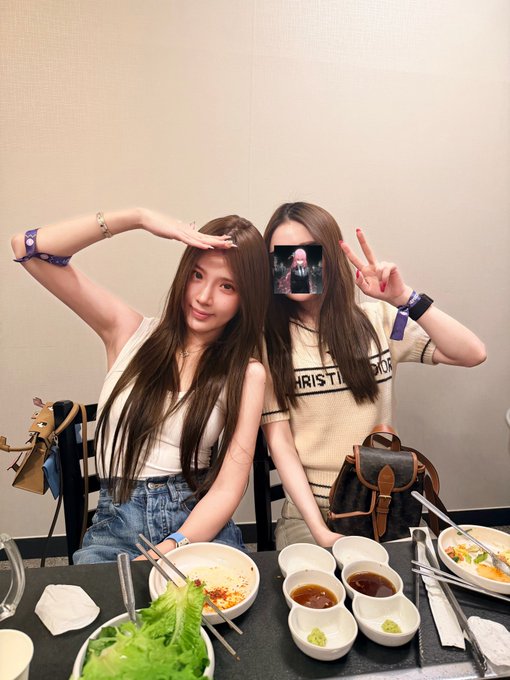 Two young women posing at a dining table; one makes a peace sign while the other's face is covered by a small inset photo, with various dishes spread before them.
