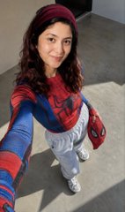 A young woman dressed in a Spider-Man costume with a red mask pulled down smiles while taking a selfie in what appears to be an outdoor setting.