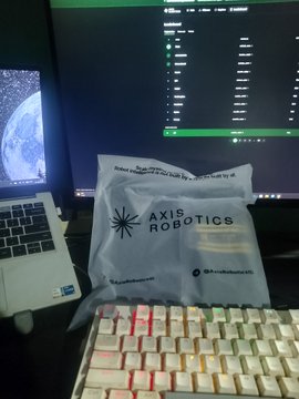 A desktop setup includes a small padded envelope branded with 'AXIS ROBOTICS' resting in front of a monitor displaying a dark-themed interface, with a white and red keyboard in the foreground and a partial view of Earth on another screen.