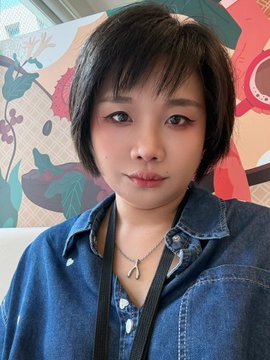 A close-up portrait of a young woman with short dark hair wearing a blue denim shirt and a silver pendant necklace, posing in front of a colorful floral wallpaper.