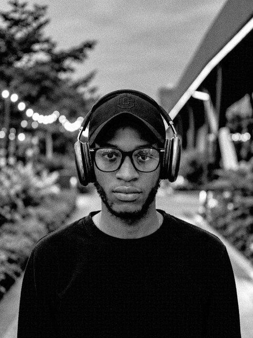 A black-and-white photo of a young man wearing glasses, a cap, and over-ear headphones, looking directly at the camera.
