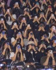 A large crowd of fans sits in stadium seats holding smartphones, many apparently recording or taking photos. The post’s joke, “Cheese… but it’s all about priorities,” reflects their focus on capturing the moment instead of watching the event.