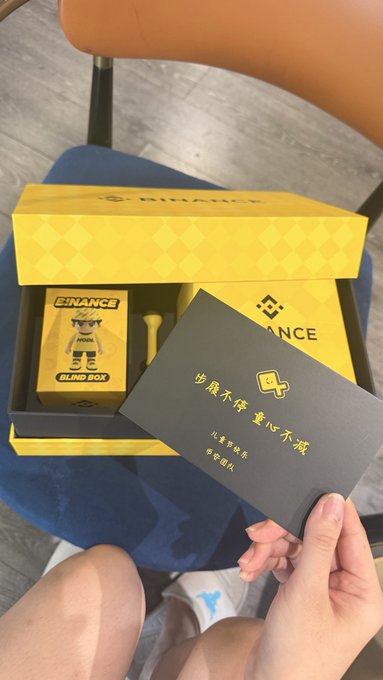 The image features a person holding up a card with Binance written on it, along with a toy robot figurine inside a box. The person's hand is visible as they hold the card and the toy. The background of the image shows a chair and a blue floor, creating an indoor setting for this scene.