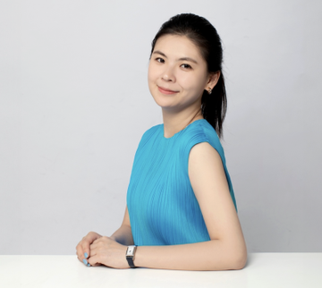 A young woman with long dark hair, wearing a blue sleeveless top and a wristwatch, smiles warmly while leaning on a white surface against a light background. The image likely represents a crypto market commentator or Bitget executive sharing bullish insights on Bitcoin's rising market dominance and the decline of low-utility altcoins.